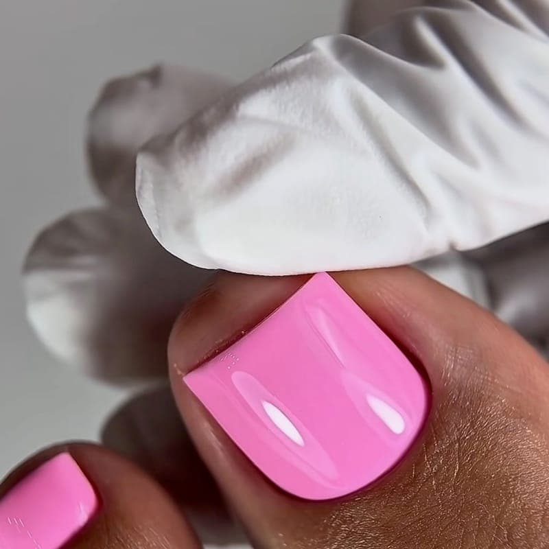 A pedicure in a bright pink gloss, a gloved hand holding a cloth beside the toe