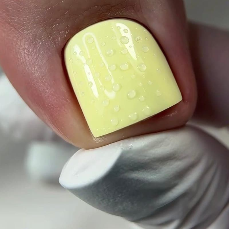 A big toe in a pale lemon gloss with water droplets, held on a white glove